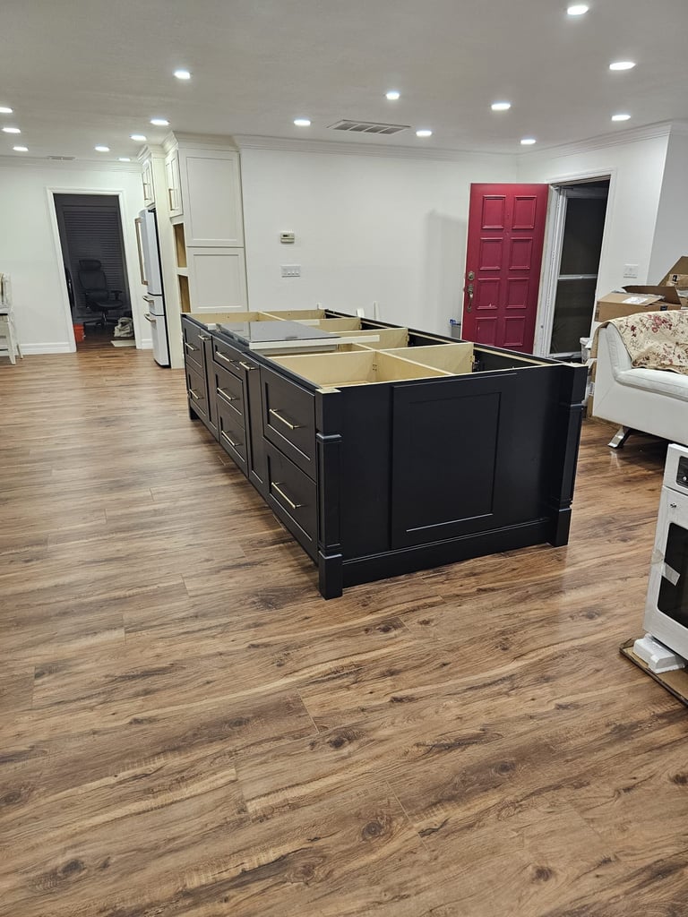 Kitchen remodel in progress with a large dark shaker-style island, white cabinetry and wood-look flooring.