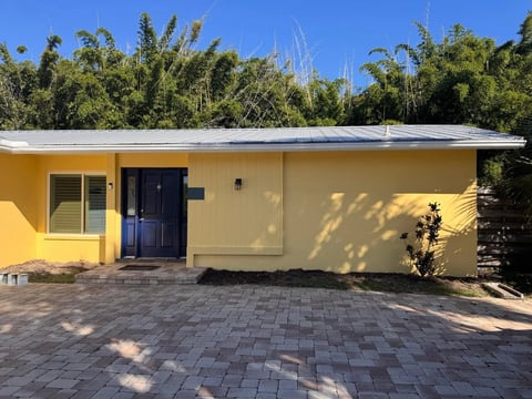 After: Finished yellow front elevation with vertical paneling, a dark blue entry door and a paver courtyard.