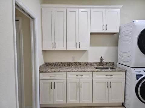 Laundry room with white cabinetry, granite countertop, sink and stacked washer and dryer units.