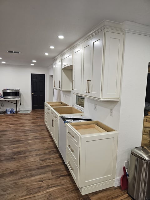 White shaker-style kitchen cabinetry with crown molding and brass-toned hardware during installation.