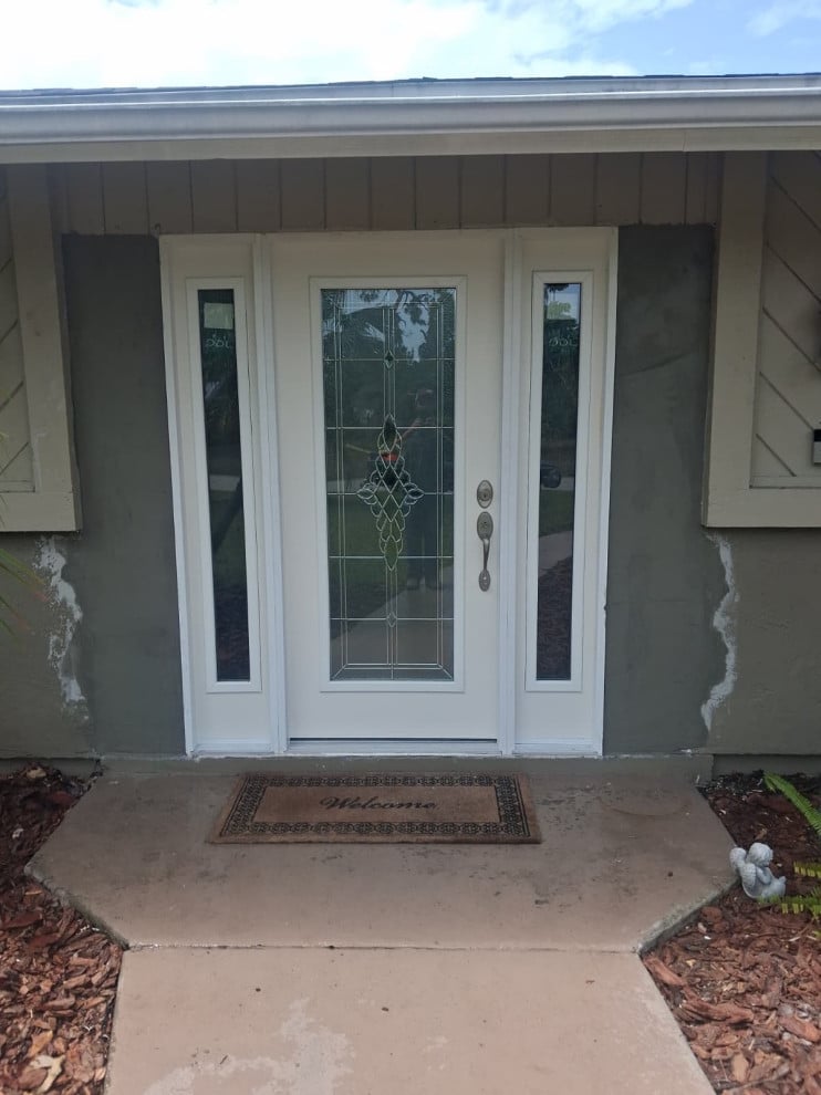 Installed white decorative-glass entry door and sidelights with patched stucco awaiting paint.