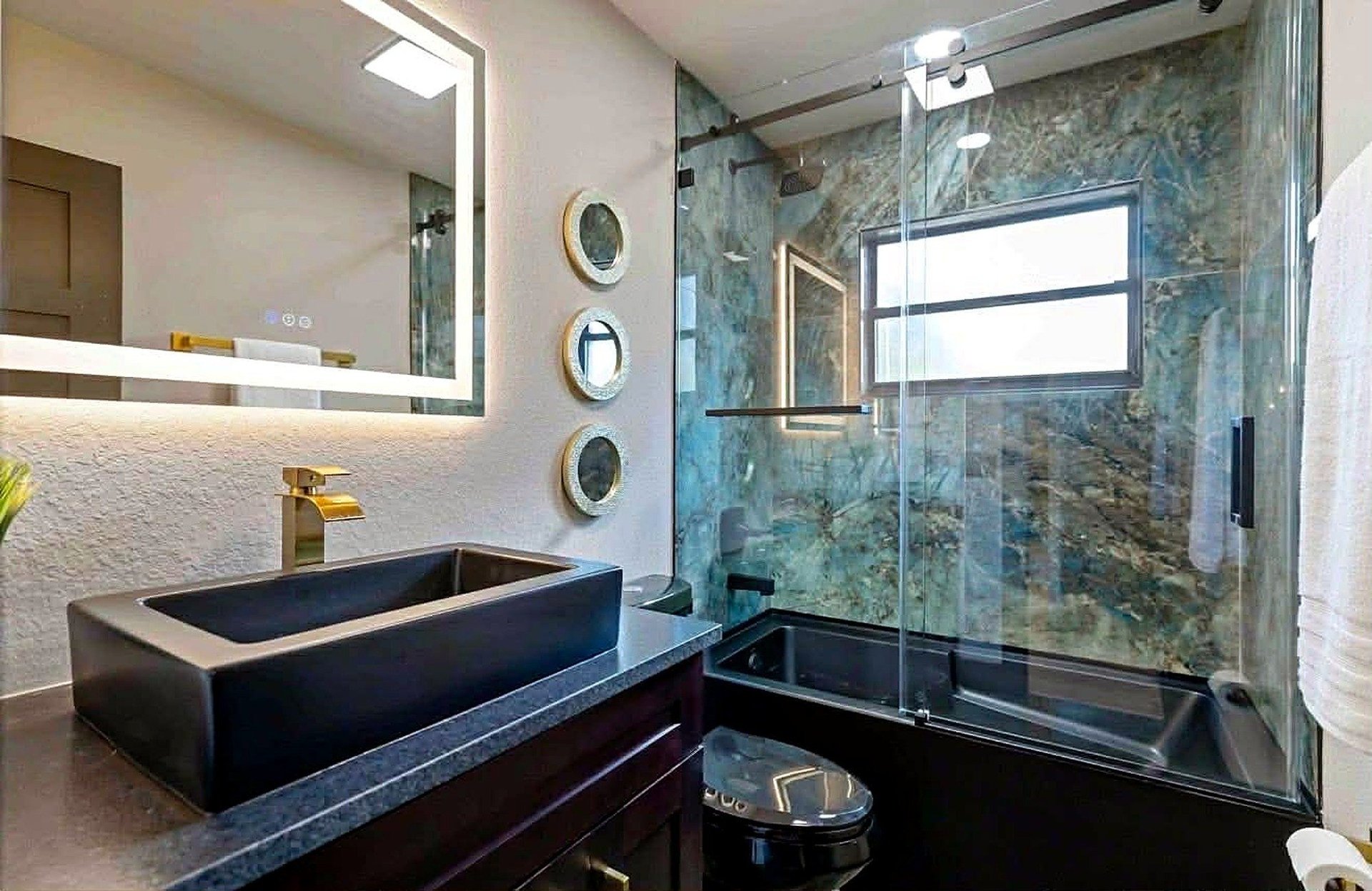 Sarasota bathroom remodel with a dark rectangular vessel sink, an illuminated mirror and blue-green tub and shower surround